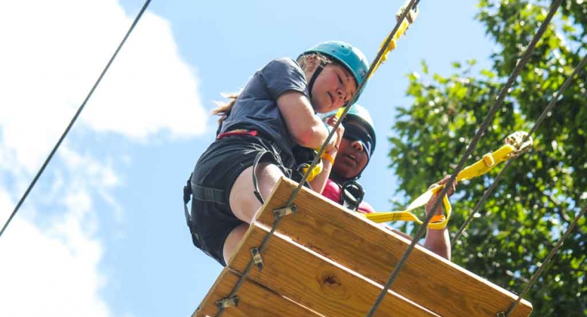 Two people wearing safety gear are attached to ropes as they navigate a high ropes course. 